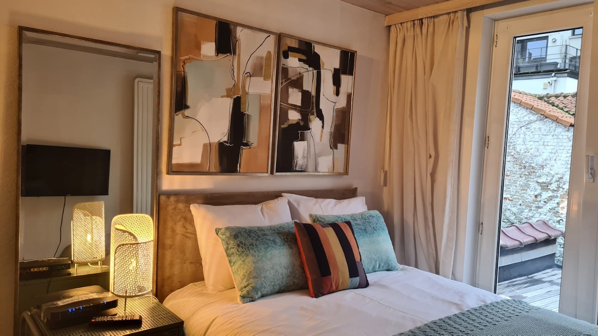 A cozy sleeping area is featured, with a spacious bed adorned by a collection of decorative pillows. Two abstract art pieces are displayed above the headboard, while a small table lamp provides warm light. A window offers a view of the outdoor space.