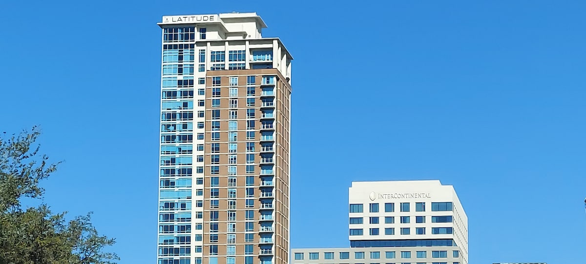 The exterior of a tall, modern building is visible against a clear blue sky. The structure features a blend of glass and brick, with multiple balconies on the upper floors. The building is distinguished by its rooftop signage and is adjacent to another prominent structure.