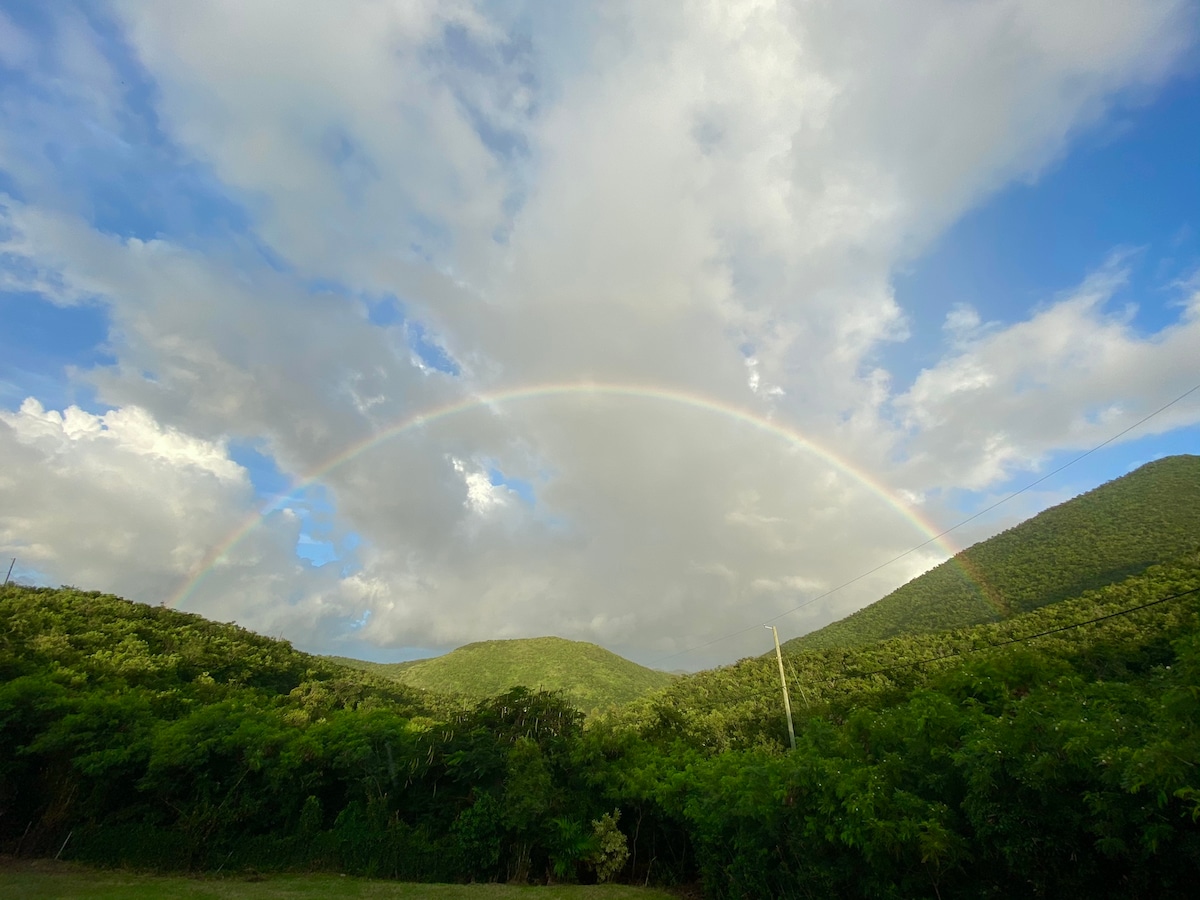 A serene landscape depicts rolling green hills beneath a dynamic sky filled with soft clouds. An arched rainbow stretches across the horizon, enhancing the natural beauty of the scene.