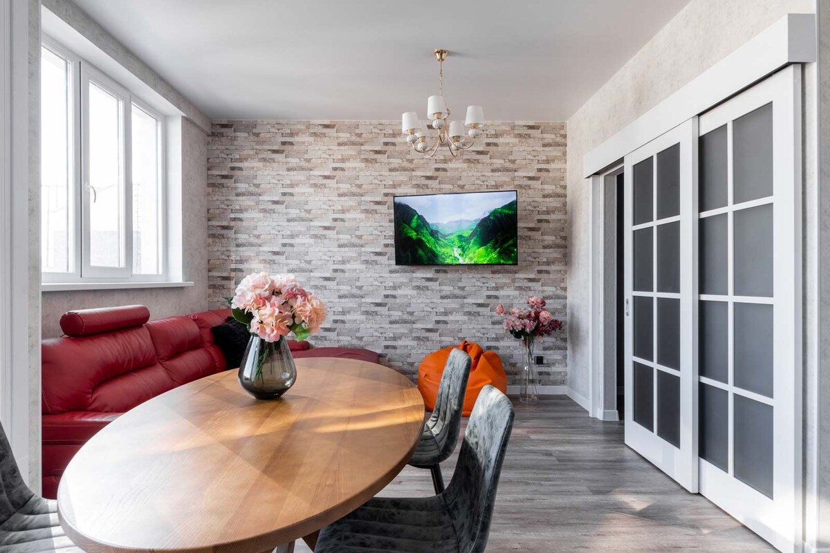 A modern living space features a round wooden dining table surrounded by chairs. A red sectional sofa offers comfortable seating, while a television displays a scenic landscape. Large windows allow natural light to flow in, and decorative elements include a vase of flowers and potted plants.