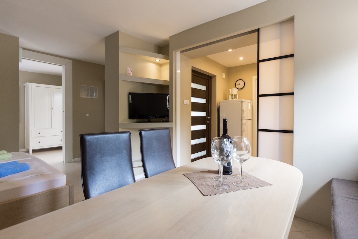 A dining area features a light-colored table set with two glasses and a bottle of wine. Dark chairs provide contrast to the neutral-toned walls. In the background, a television and refrigerator are visible, along with an open doorway leading to another room.