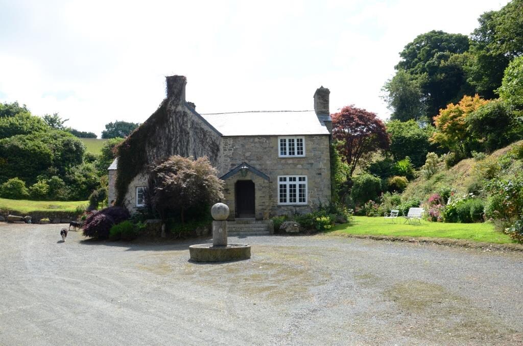 The stone exterior of a charming cottage is visible, enveloped by lush greenery and colorful shrubs. A circular stone feature stands prominently in the gravel driveway, inviting guests to explore the peaceful surroundings of the property.