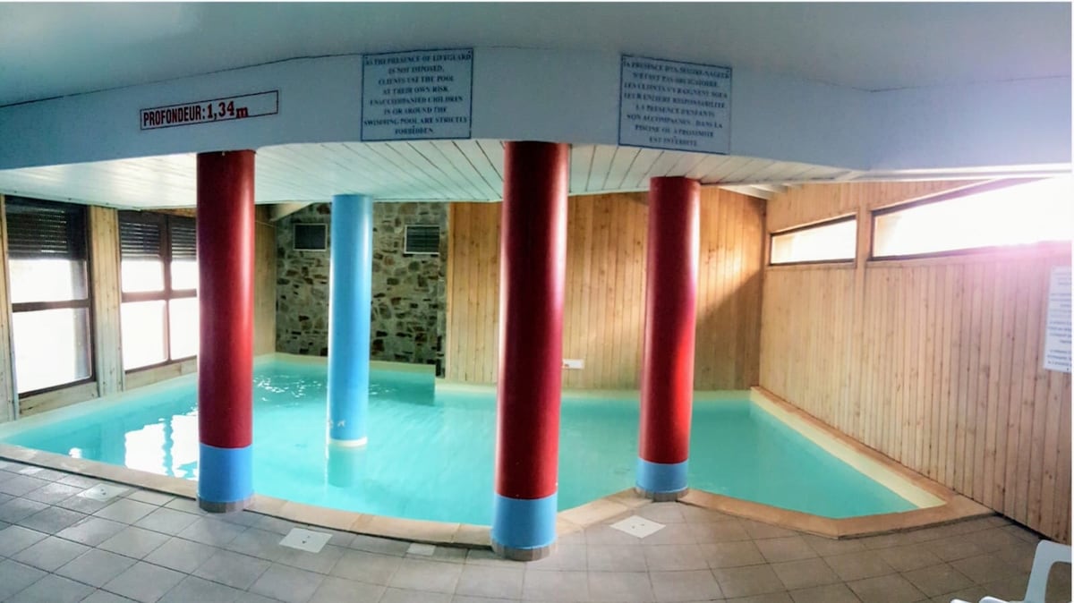 An indoor heated pool features clear turquoise water, surrounded by wooden paneling and stone accents. Four colorful columns in red and blue support the ceiling. Large windows allow natural light to fill the space, reflecting off the calm surface of the water.
