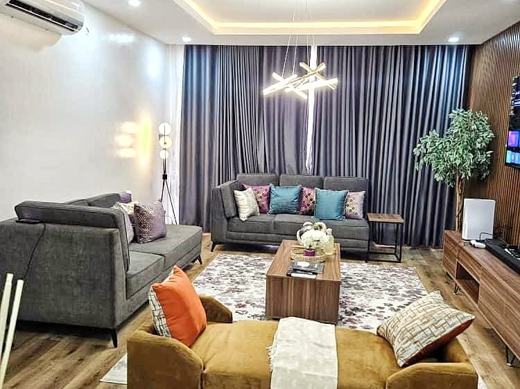 A modern living room features two gray sofas with colorful cushions, complemented by a cozy golden chair. A wooden coffee table occupies the center, adorned with a floral arrangement. Ambient lighting is provided by a unique chandelier, with large curtains framing the windows.