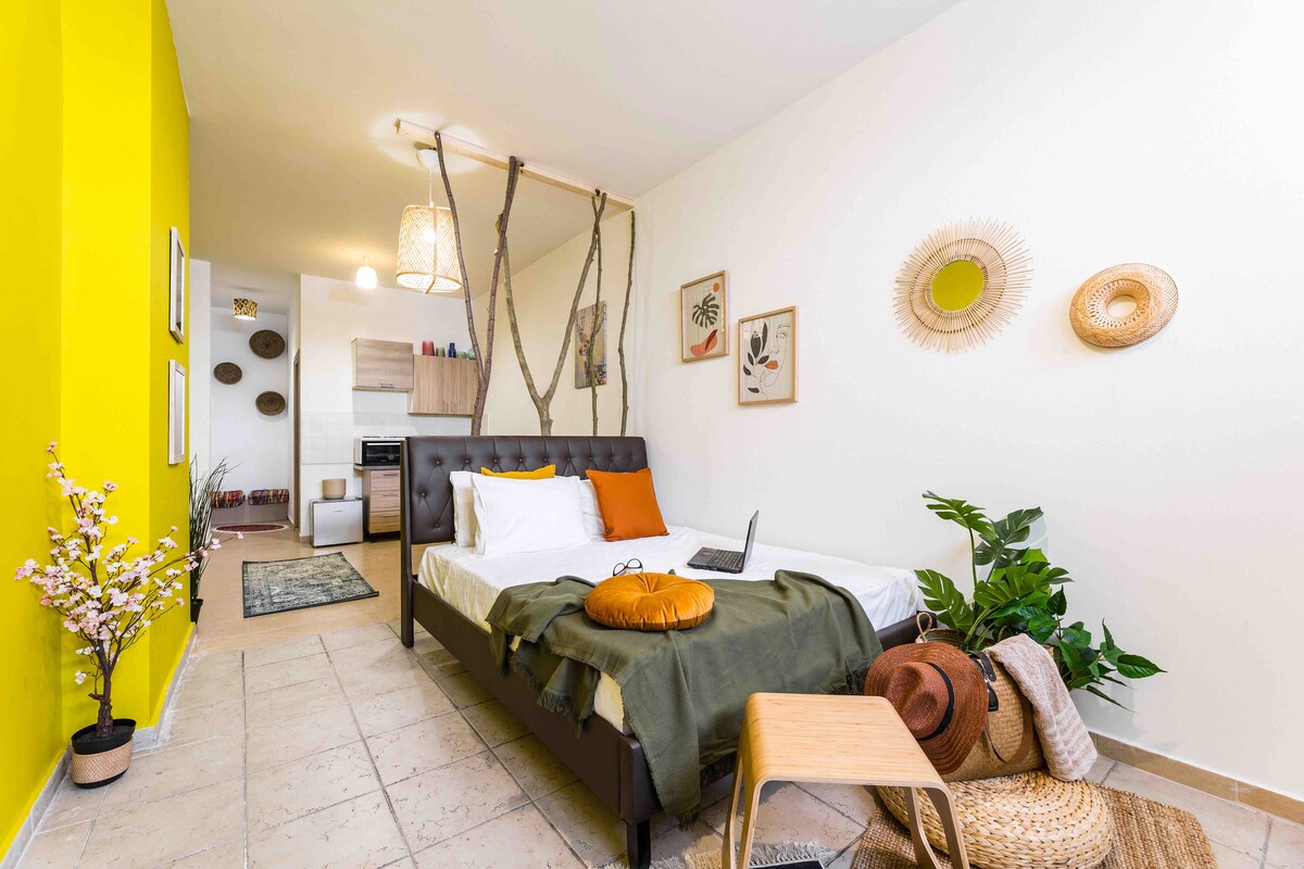 A modern studio layout reveals a spacious bed framed with a unique design. Bright yellow walls add warmth, complemented by decorative wall art. A small table and natural elements such as plants and woven baskets enhance the space's inviting feel.