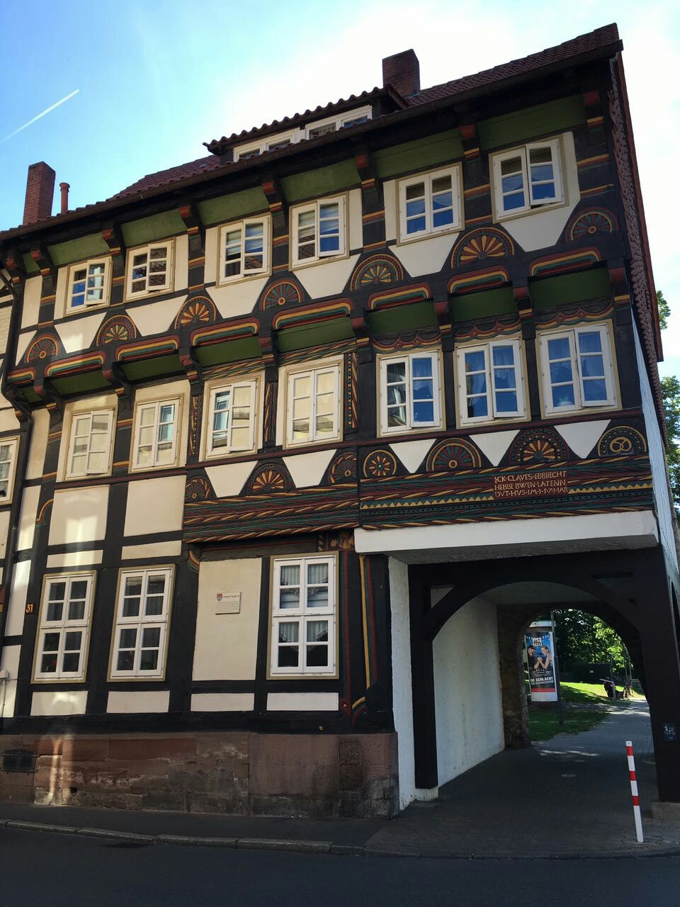 A historic building features a striking half-timbered facade adorned with colorful decorative patterns. Multiple windows line the upper levels, allowing natural light to fill the interiors. An arched entrance at the lower level provides access, contributing to the building's architectural charm.