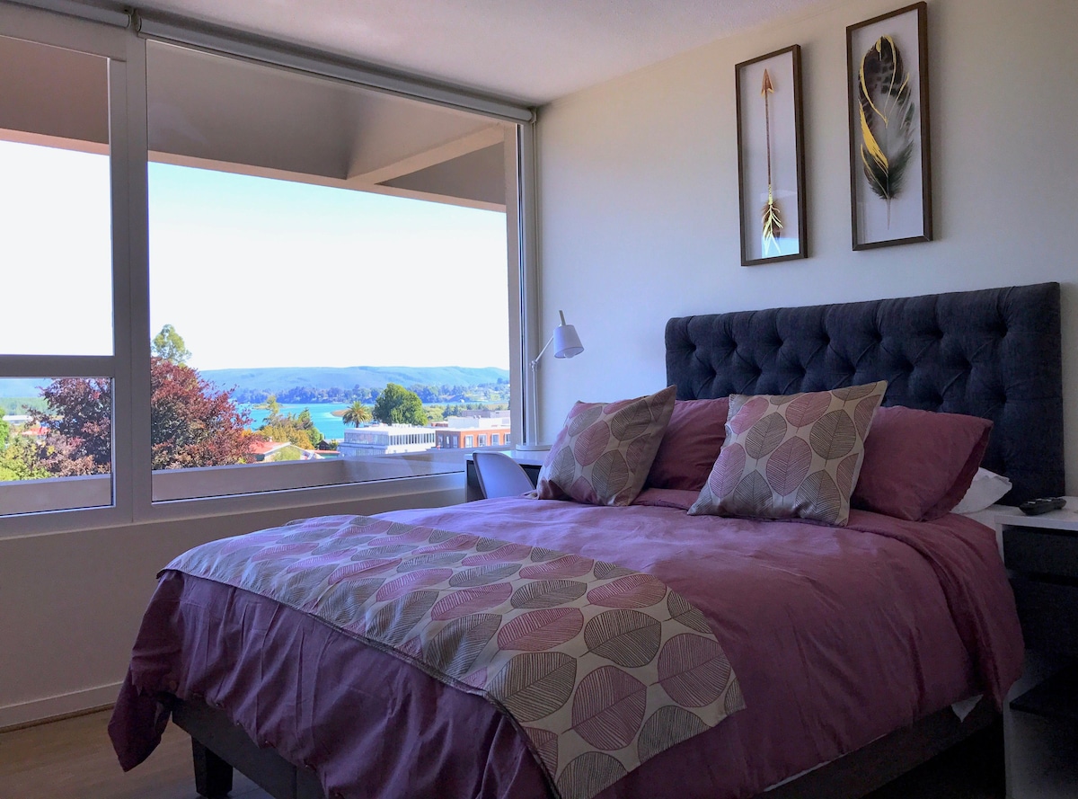 A spacious bed is adorned with a light purple bedspread and decorative pillows. Large windows frame a view of the river and surrounding hills, allowing natural light to fill the room. Framed artwork complements the neutral tones of the decor.