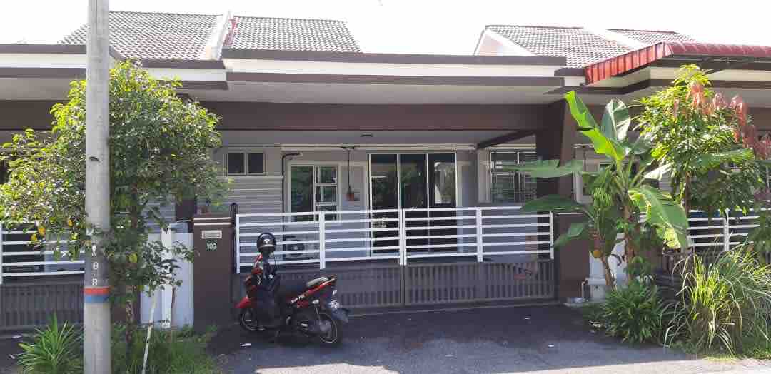 The exterior of the homestay is displayed, showcasing a modern single-story structure with a covered porch. A motorcycle is parked in front, and green plants frame the entrance. Large windows allow natural light to enter the interior space.