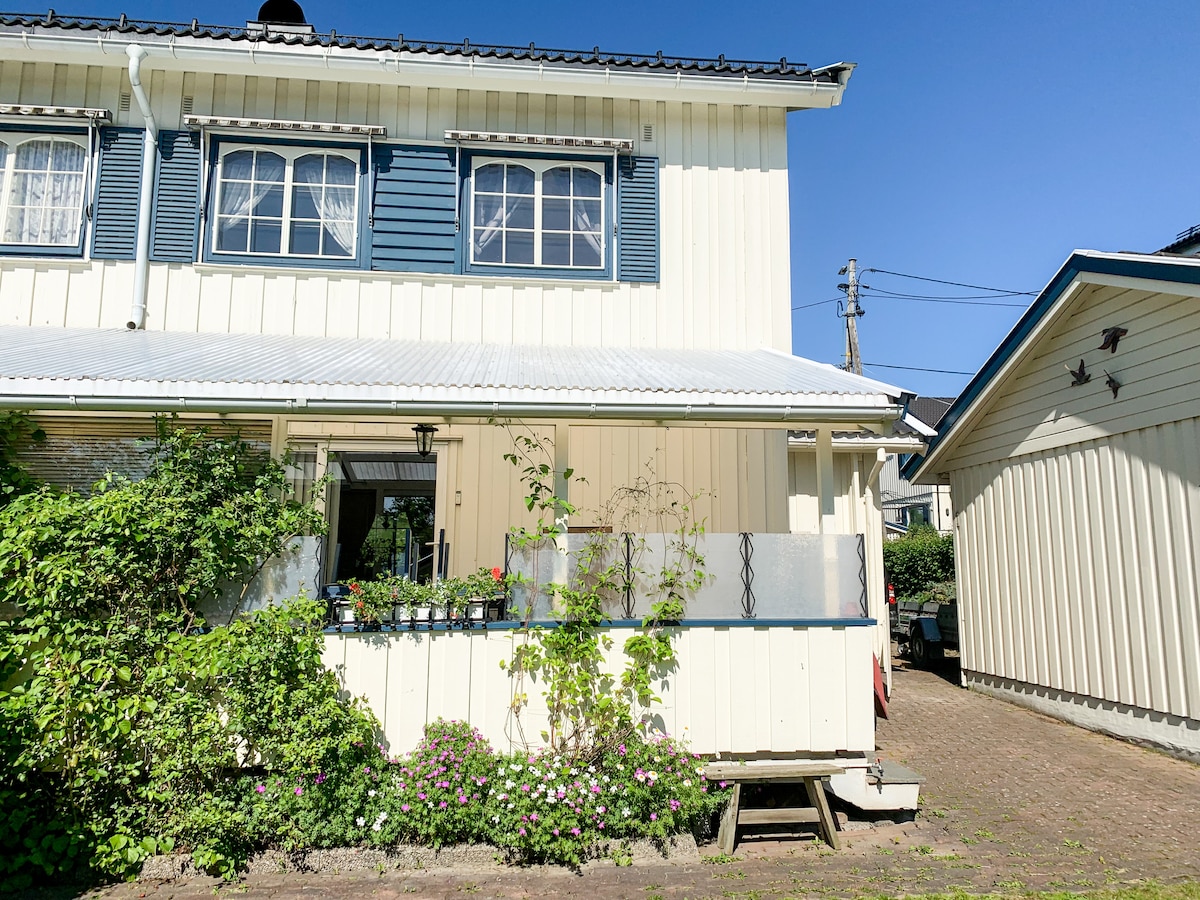 The exterior of a two-family home is displayed, featuring a combination of light-colored and blue siding. A covered porch is seen with various potted plants, and colorful flowering vines are growing alongside. Bright blue skies enhance the inviting atmosphere.