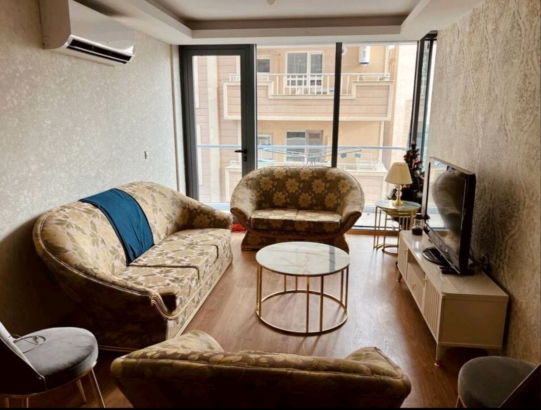 A cozy living area is presented, featuring a floral sofa set and a round marble coffee table. Large glass windows allow natural light to fill the space, showcasing a view of the balcony. A television is positioned on a low cabinet, enhancing the functional design.