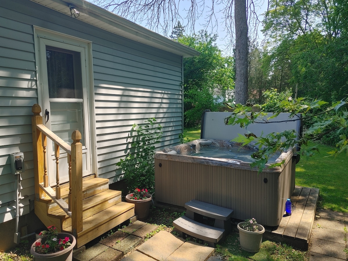 A hot tub is situated on a wooden deck beside a light gray house, surrounded by lush greenery. Flower pots with vibrant blooms add color to the scene. The entrance to the house is accessible via a small staircase.