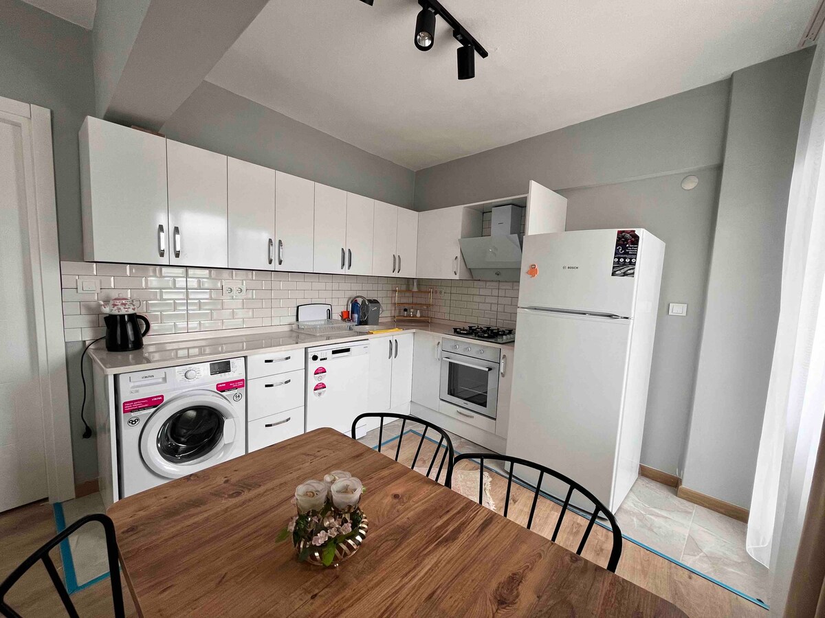 A modern kitchen features white cabinets and essential appliances, including a refrigerator, stove, and washing machine. A wooden table with black chairs is positioned in the dining area, with a small floral centerpiece enhancing the space.