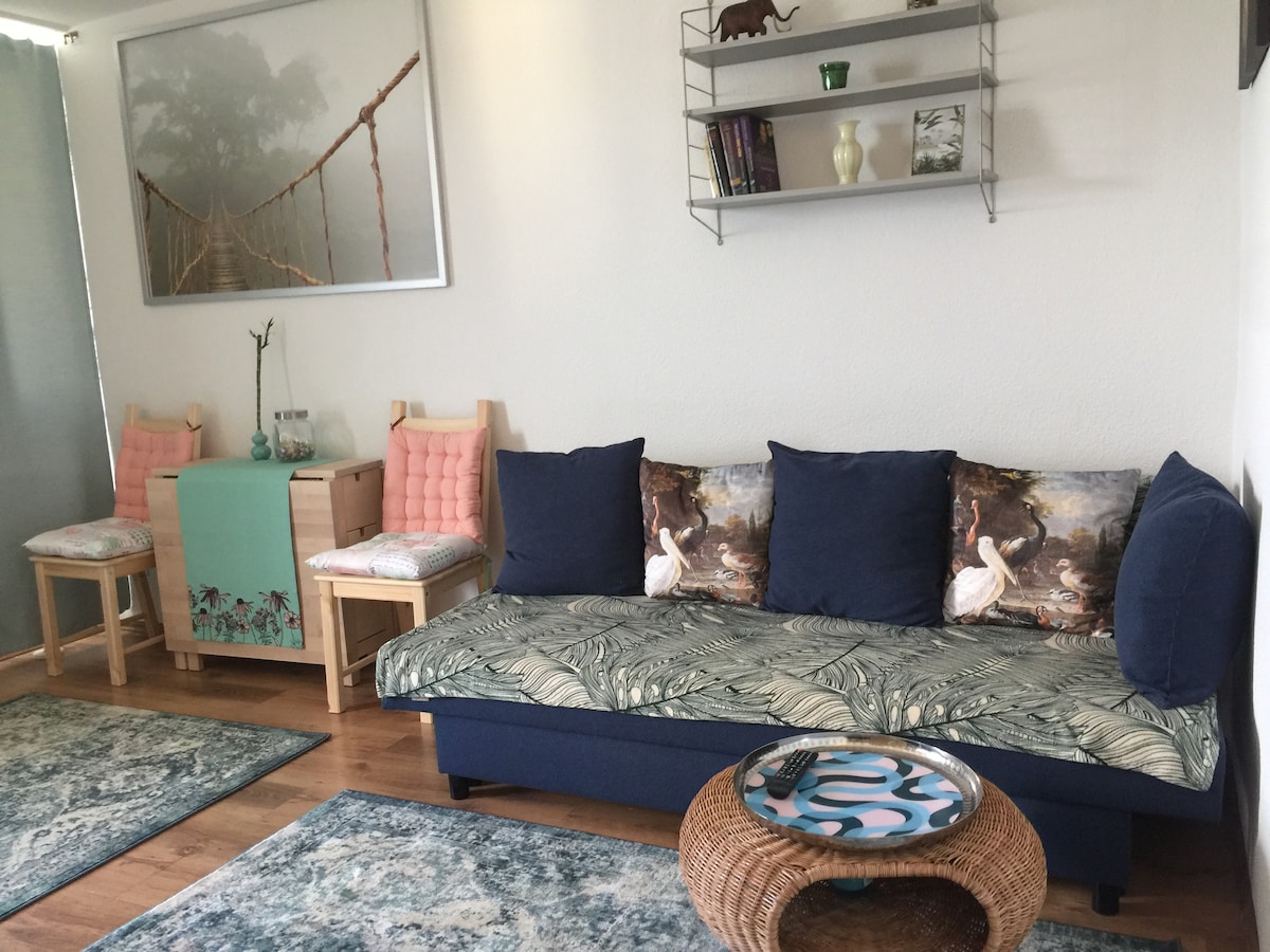 A cozy living area is showcased, featuring a spacious blue sofa adorned with patterned cushions. The hardwood floor is complemented by area rugs, while a round woven coffee table adds a natural touch. Shelves with decorative items provide additional visual interest.