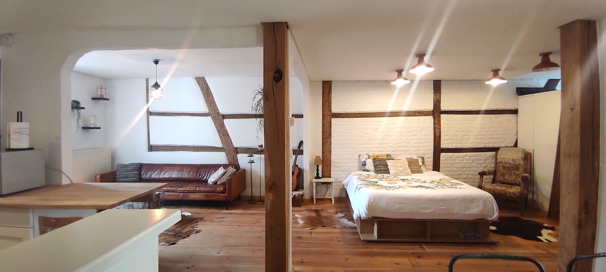 An open living space features a cozy sleeping area with a spacious bed dressed in a colorful patchwork quilt. A leather sofa and rustic wooden beams add character, while warm lighting creates a relaxed atmosphere. A small table and chair complement the inviting layout.