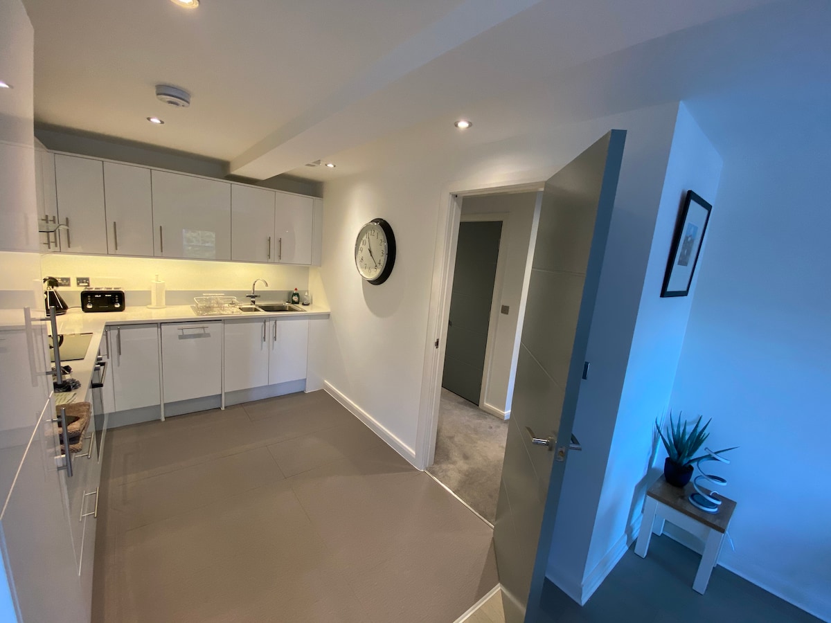 The kitchen area features modern cabinetry and appliances, including a microwave and induction hob. Natural light fills the space, highlighting the clean lines and contemporary design. A large round clock is mounted on the wall, complementing the overall layout.