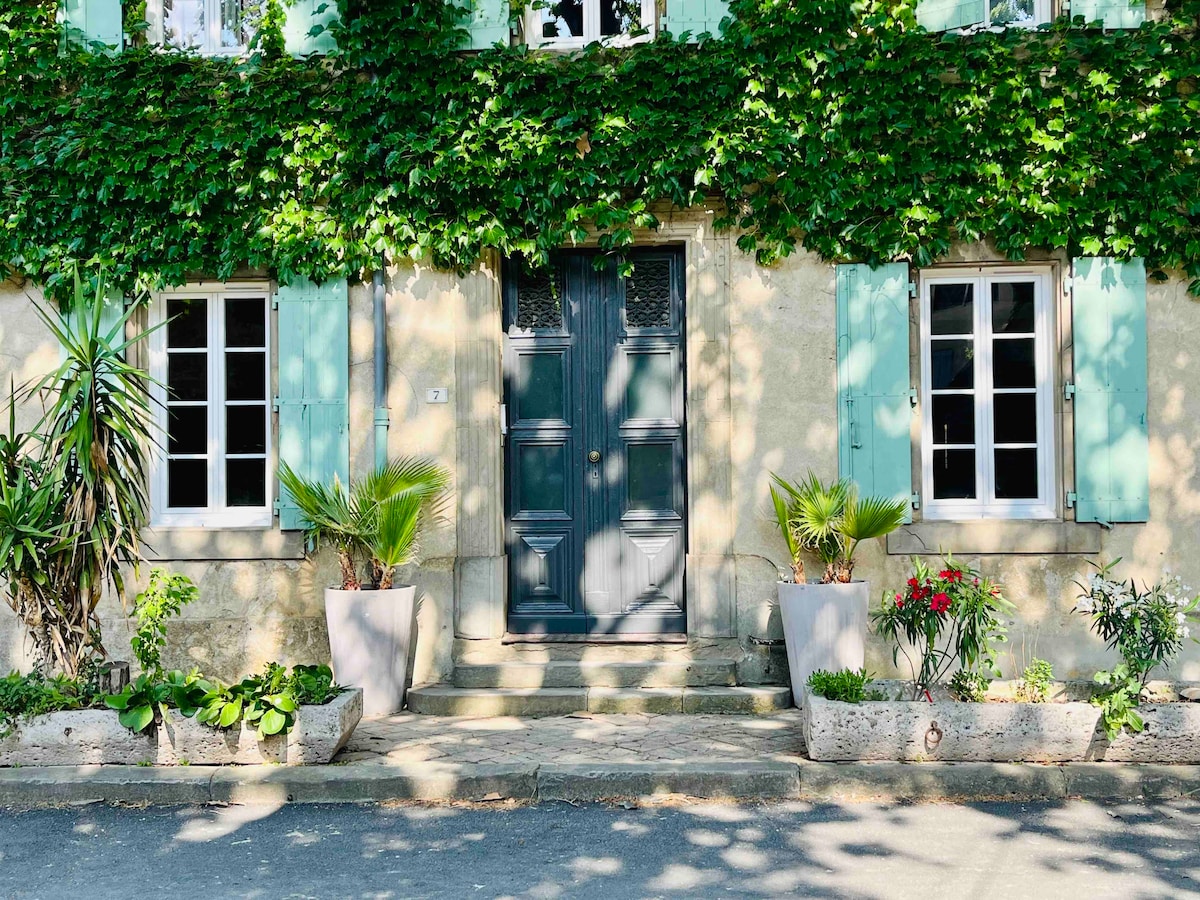 The exterior of a charming residence is framed by climbing greenery and features light green window shutters. Two large planters with a variety of plants and flowers are placed on either side of a set of stone steps leading to a muted blue entrance door.