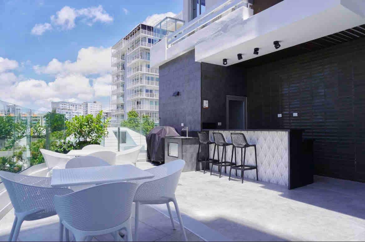 An outdoor terrace is featured with a modern bar area and four white chairs surrounding a circular dining table. The backdrop includes greenery and contemporary buildings under a bright blue sky, enhancing the spacious feel of the space.