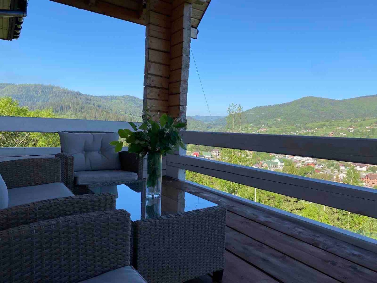 A covered terrace features comfortable seating and a glass table, with a scenic view of the mountains and valley. Lush greenery is visible in the background, providing a peaceful ambiance for relaxation.