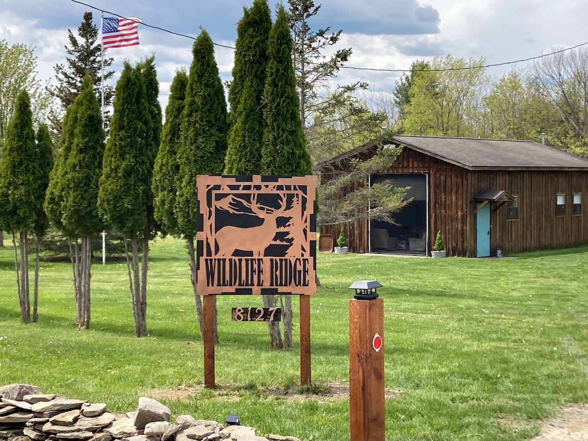 A wooden sign displaying 'WILDLIFE RIDGE' stands in front of a cabin surrounded by neatly trimmed greenery. A flag fluttering in the breeze is visible nearby. The cabin structure features a combination of wood siding and large windows, adding to the rustic charm of the setting.