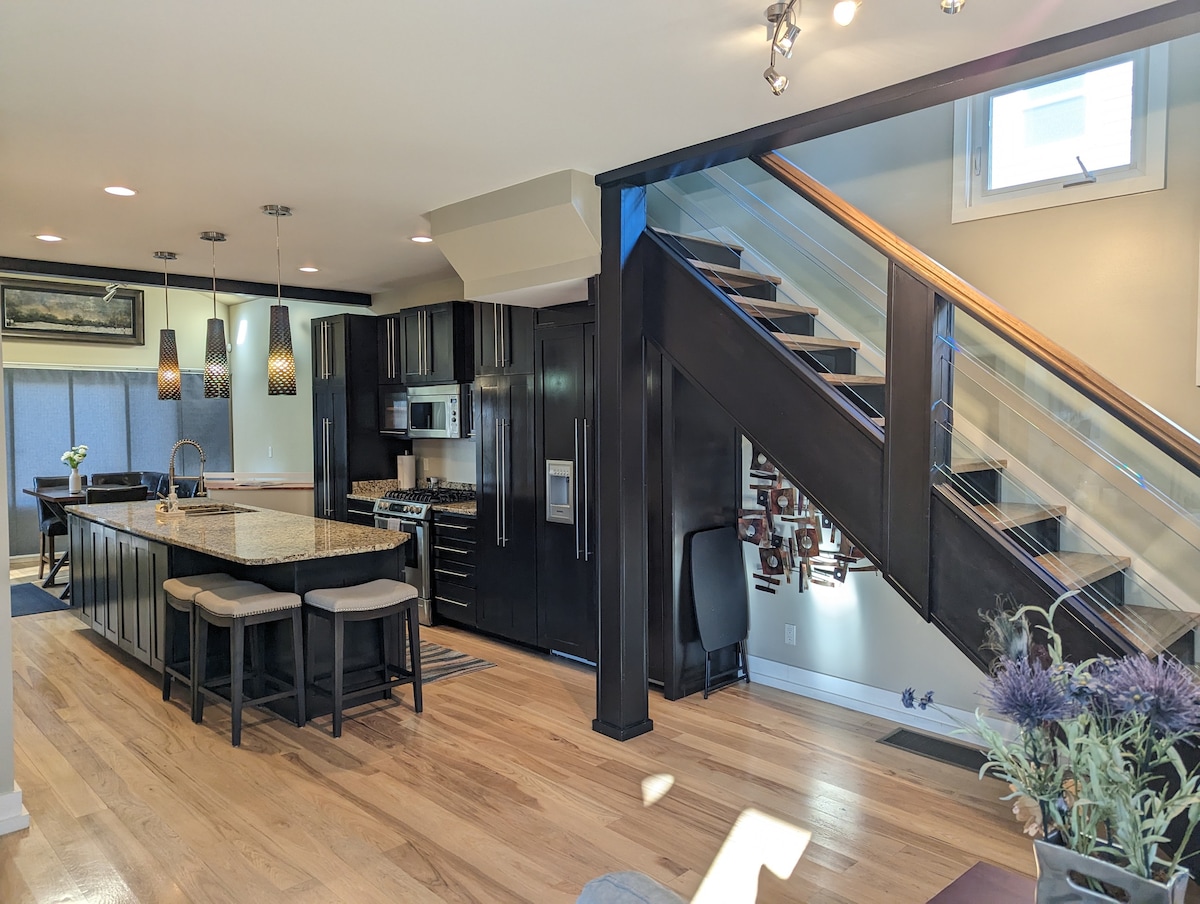 The modern kitchen features a large island with seating, sleek cabinetry, and stainless-steel appliances. A staircase with glass railings leads to the upper level. Pendant lights illuminate the workspace, while natural light filters through the windows, enhancing the inviting atmosphere.