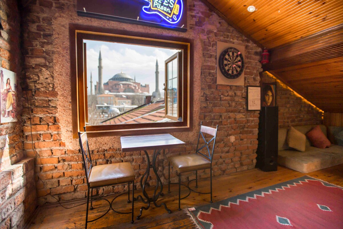 A cozy corner features a small table with two metal chairs, set against a backdrop of exposed brick walls. A window offers a scenic view of Hagia Sophia. The warm wooden ceiling and colorful rug enhance the inviting atmosphere of the space.