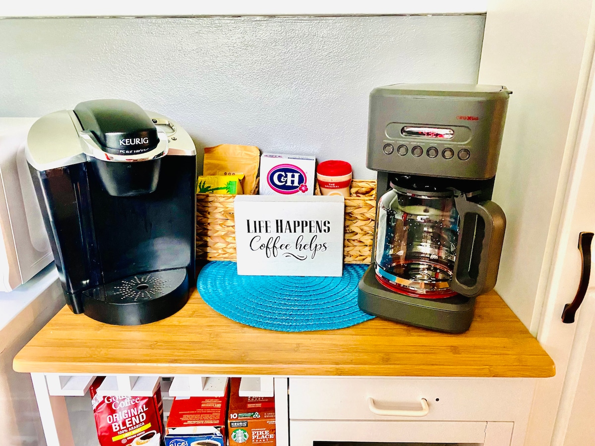 A coffee station is displayed, featuring a single-serve brewing machine alongside a traditional coffee maker. A small sign reads, 'Life happens, coffee helps.' A woven basket contains various coffee supplies, all set on a wooden countertop accented by a blue circular mat.