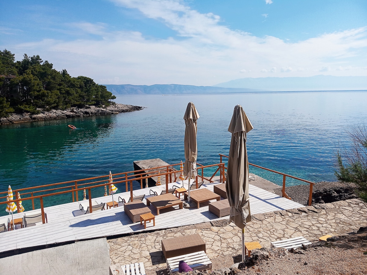 A serene outdoor space is featured, showcasing a wooden deck with sun loungers and umbrellas overlooking calm, clear waters. Lush trees are visible in the background, while distant hills can be seen across the water, enhancing the peaceful ambiance of the area.