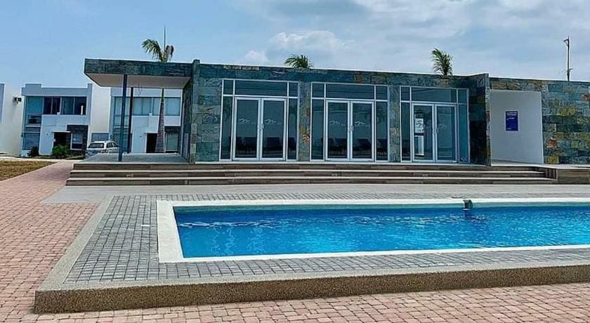 A modern house is shown with large glass doors and a sleek stone façade. A rectangular swimming pool sits in the foreground, surrounded by a wide stone patio. The area is spacious and features palm trees in the background under a clear sky.