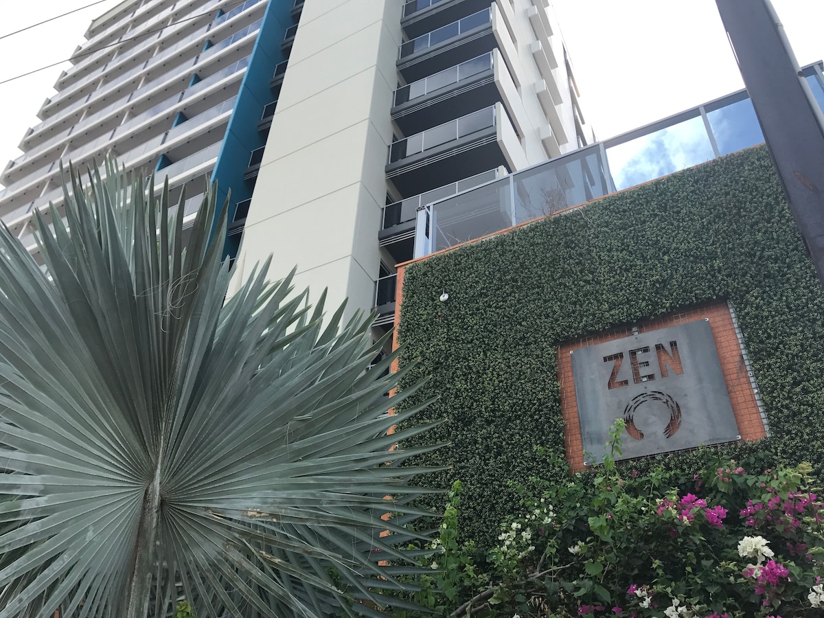 The exterior of the Ramada Zen Quarter building is showcased, featuring a green wall adorned with foliage and a decorative sign reading 'ZEN.' A tall palm plant is positioned in the foreground, complementing the modern architectural design of the building.
