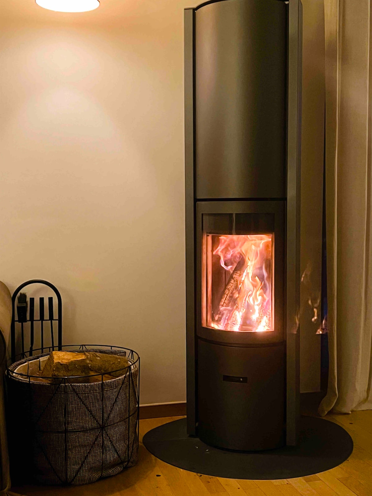 A modern wood stove stands in the corner of a cozy living room, with flames visible through its glass front. Next to the stove, a wire basket is filled with neatly stacked firewood, adding a functional element to the warm ambiance.