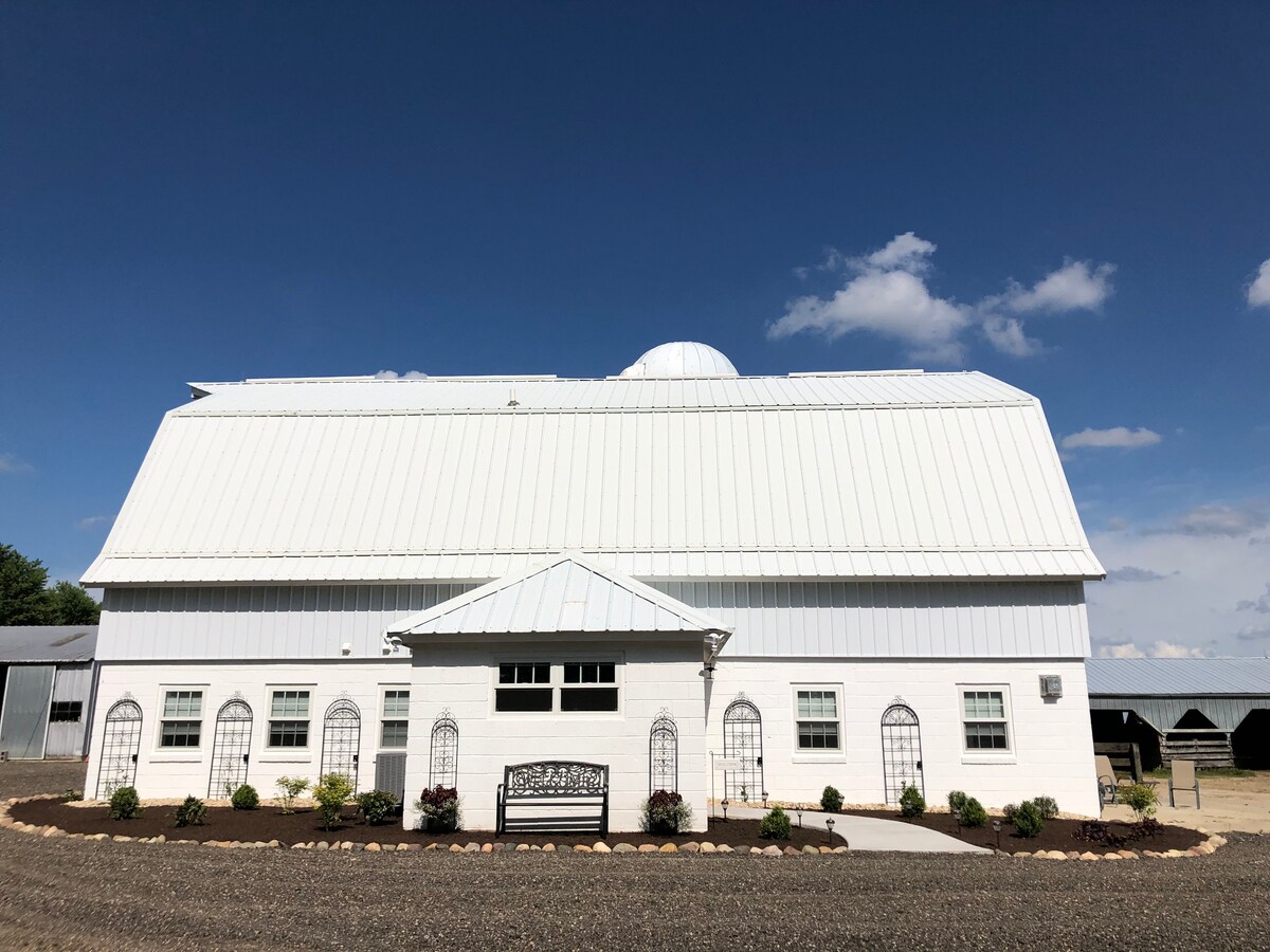 The sturdy white barn features a distinctive metal roof and multiple gabled sections. A welcoming entrance is highlighted by decorative lattice fencing and a landscaped area with plants. Clear blue skies and clouds create a calm backdrop, enhancing the building's spacious exterior.