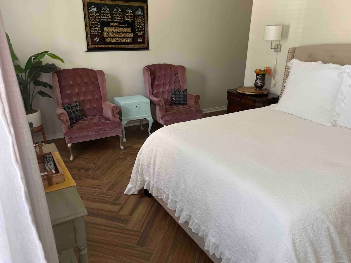 A cozy bedroom is highlighted by a spacious bed with a white coverlet, complemented by two elegant pink armchairs and a small side table. A piece of art hangs above the chairs, and natural light filters in through a window, enhancing the inviting setting.