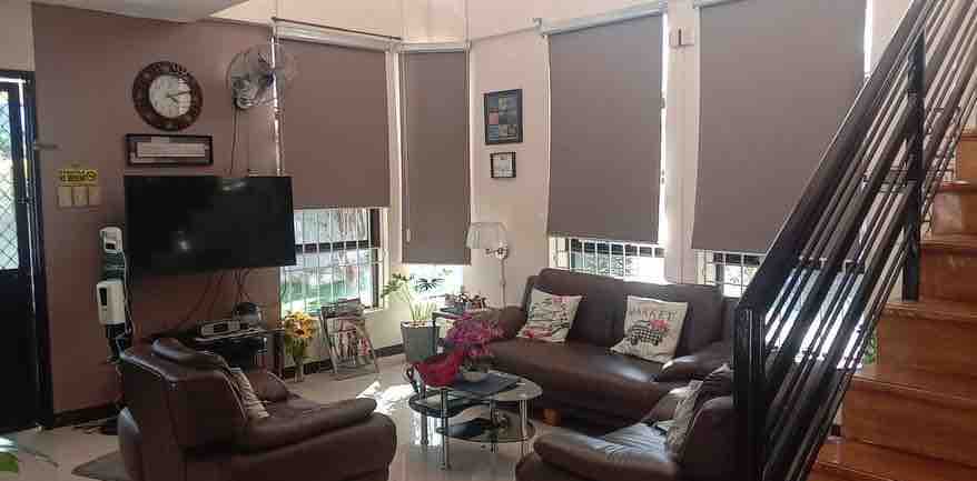 A cozy living space is depicted with a brown leather sofa and matching armchairs arranged around a small coffee table. Large windows with light-filtering shades allow natural light to fill the room, while a wall-mounted television is visible. A staircase leads to an upper level.