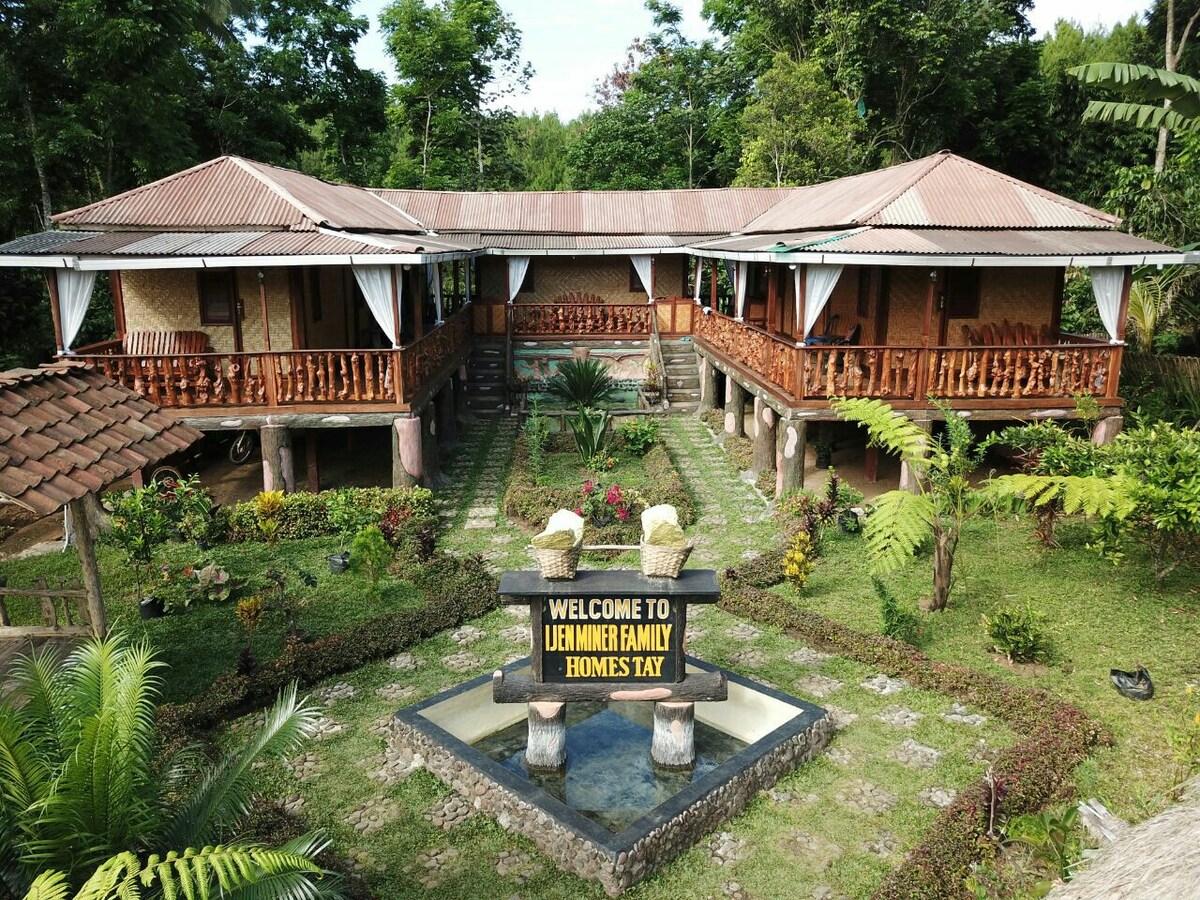A spacious outdoor area is bordered by vibrant gardens featuring a variety of plants. Two traditional wooden houses are positioned symmetrically, each with covered terraces. A welcoming sign sits in the center, surrounded by decorative stones and greenery, inviting guests to the home stay.