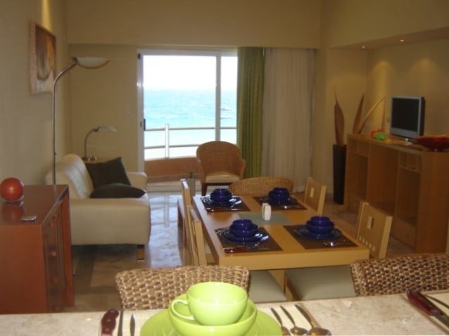 An open living space features a dining table set for six with blue dishware, complemented by a comfortable seating area. Large glass doors provide a view of the ocean and allow natural light to fill the room, highlighting the neutral color palette and modern furnishings.