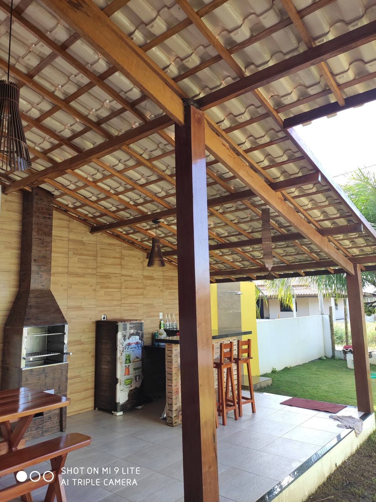 An outdoor gourmet area with a thatched roof features a barbecue grill, a small refrigerator, and a bar area with high stools. Wooden dining tables are present, and natural light filters through the structure, illuminating the stone accents and grassed area outside.