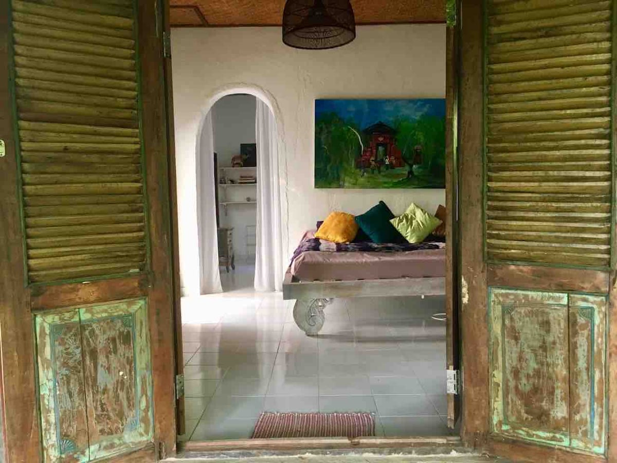 An inviting entryway with wooden shutters opens to reveal a cozy bedroom featuring a spacious bed adorned with patterned pillows. A vibrant painting hangs on the wall, complemented by soft curtains and tiled flooring, creating a bright and airy atmosphere.