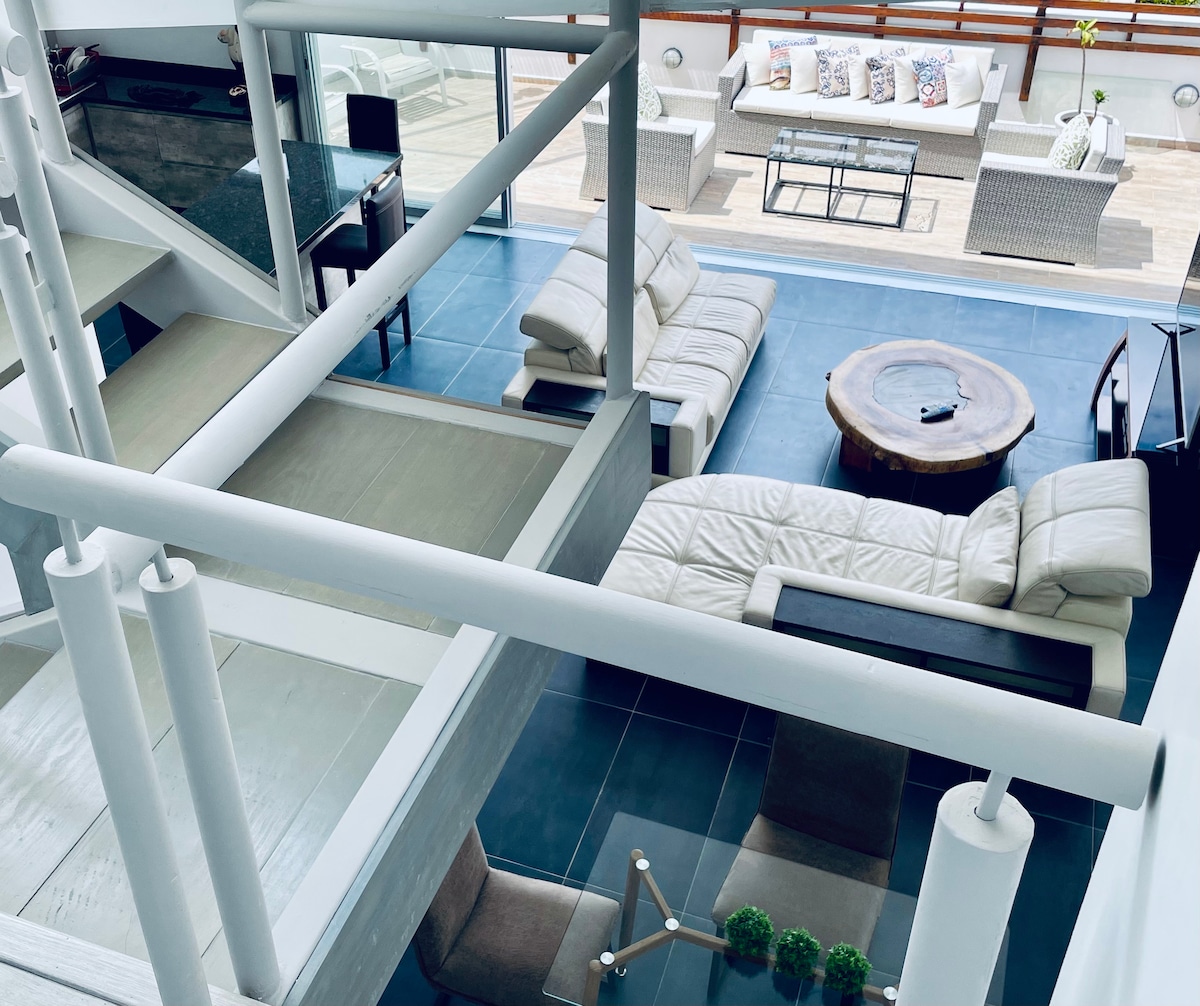 A modern living area features a spacious sectional sofa and a round wooden coffee table. Large windows allow for natural light, while a dining area with high chairs is visible in the background. A terrace with outdoor seating can be seen beyond the glass doors.