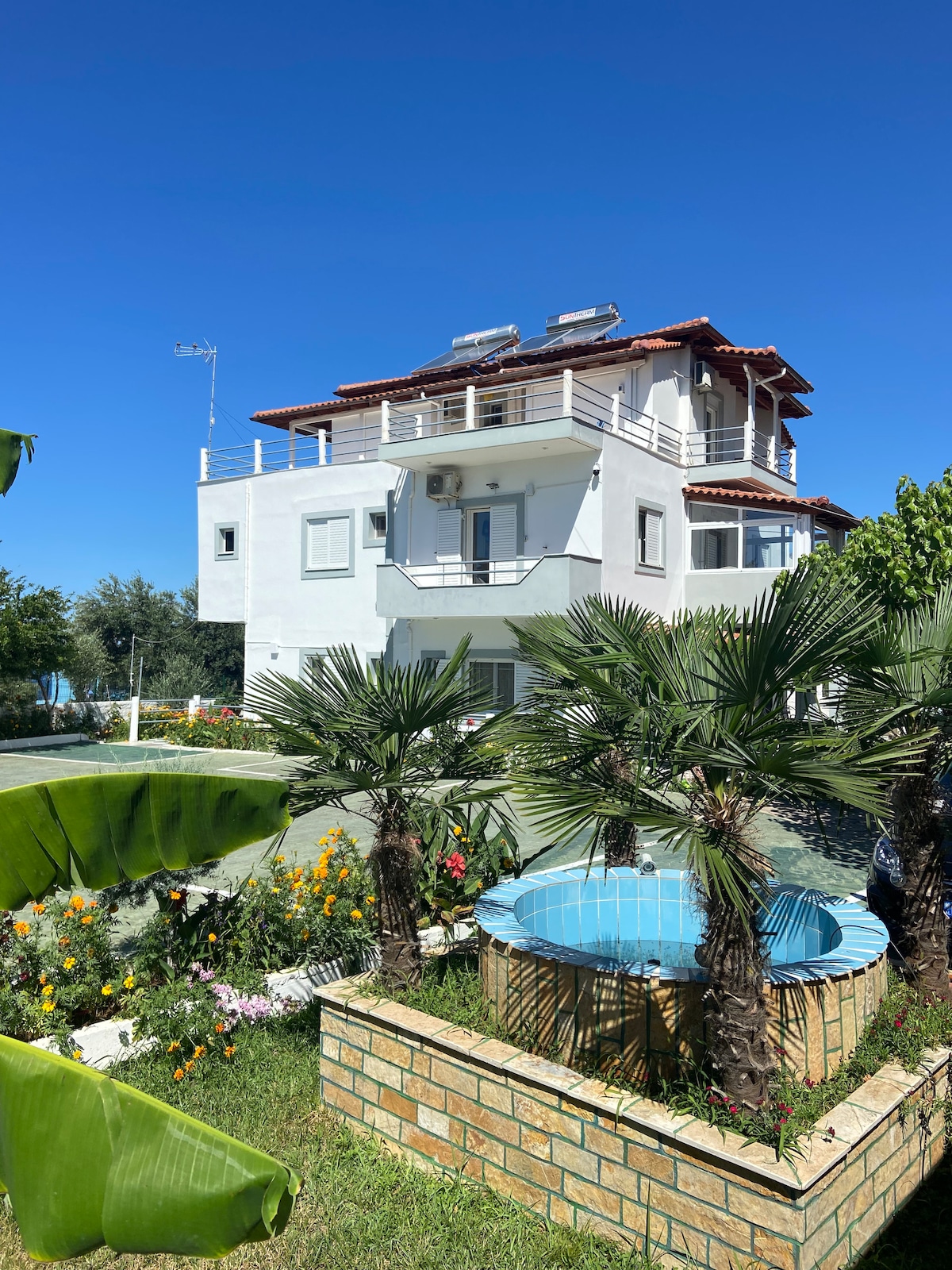 The exterior of a multi-level white building is featured, surrounded by lush greenery and colorful flowerbeds. Palm trees border a circular blue-tiled pool, adding a tropical touch. A clear blue sky enhances the inviting atmosphere of this well-maintained property.