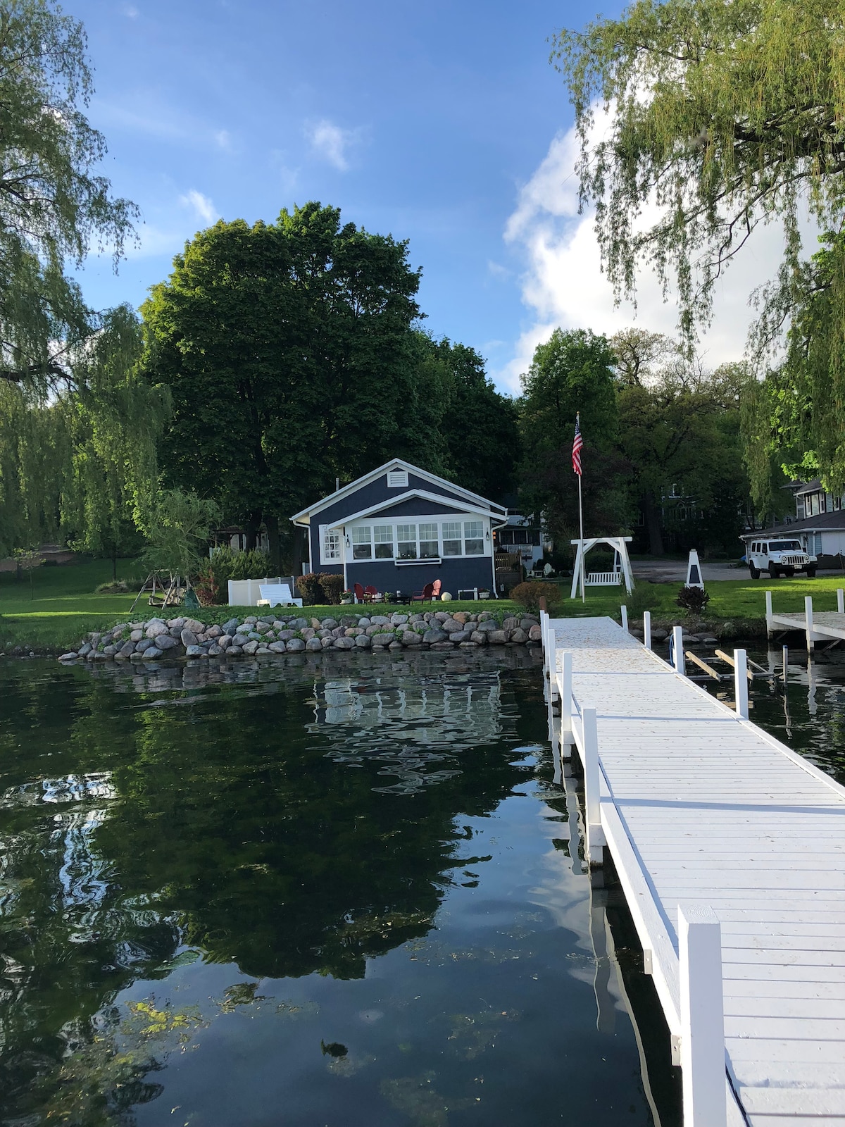 The lakefront cottage is framed by lush greenery and is complemented by a stone-lined garden. A private white pier extends into the calm waters, providing access to the lake. The exterior features large windows and a welcoming porch, with a flag gently waving nearby.