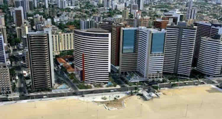 An aerial view reveals a modern urban landscape characterized by tall, sleek buildings lining a sandy beach. The coastline stretches alongside the city, showcasing a blend of natural and architectural elements amid a vibrant setting.