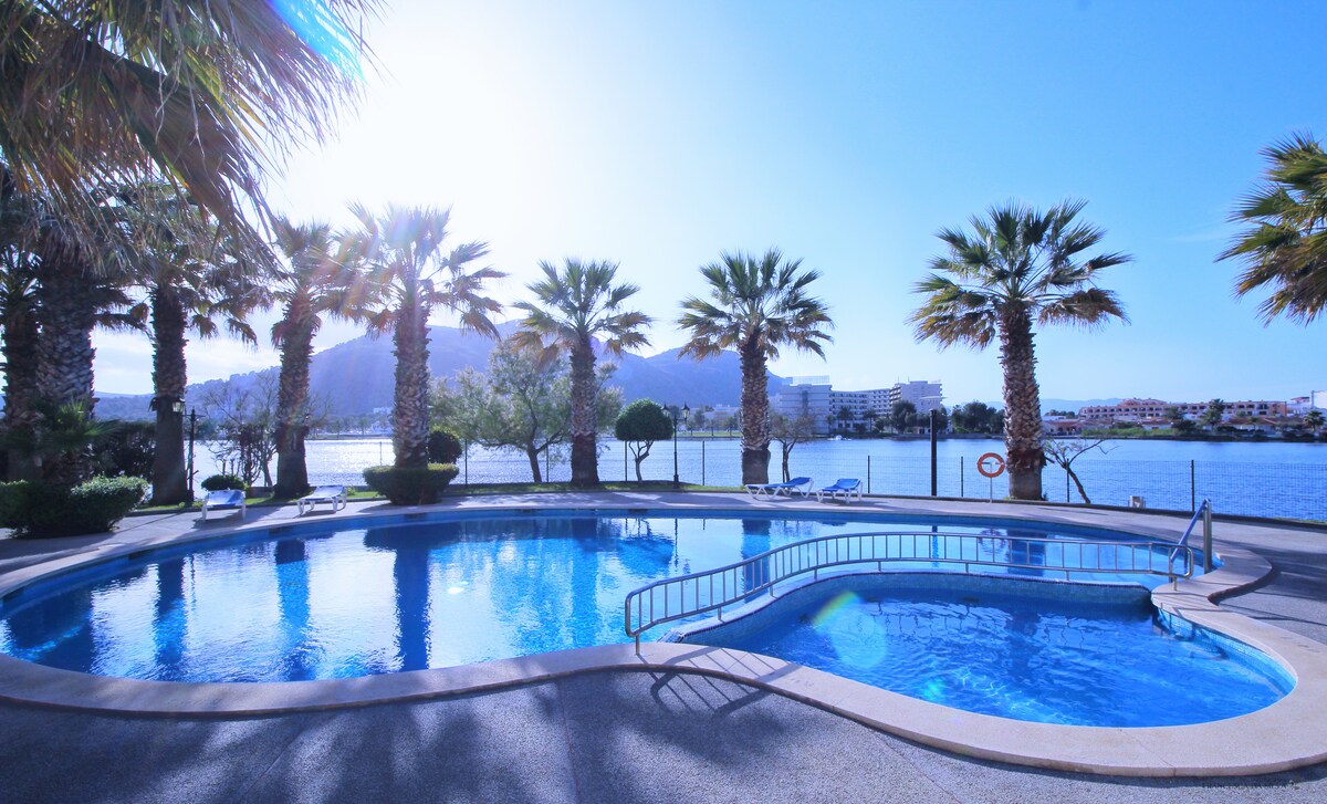 A clear swimming pool is surrounded by palm trees, offering a serene atmosphere. Sunlight reflects off the water, creating a tranquil scene. Lounge chairs are positioned nearby, and a gentle slope leads to the pool's edge, providing easy access.