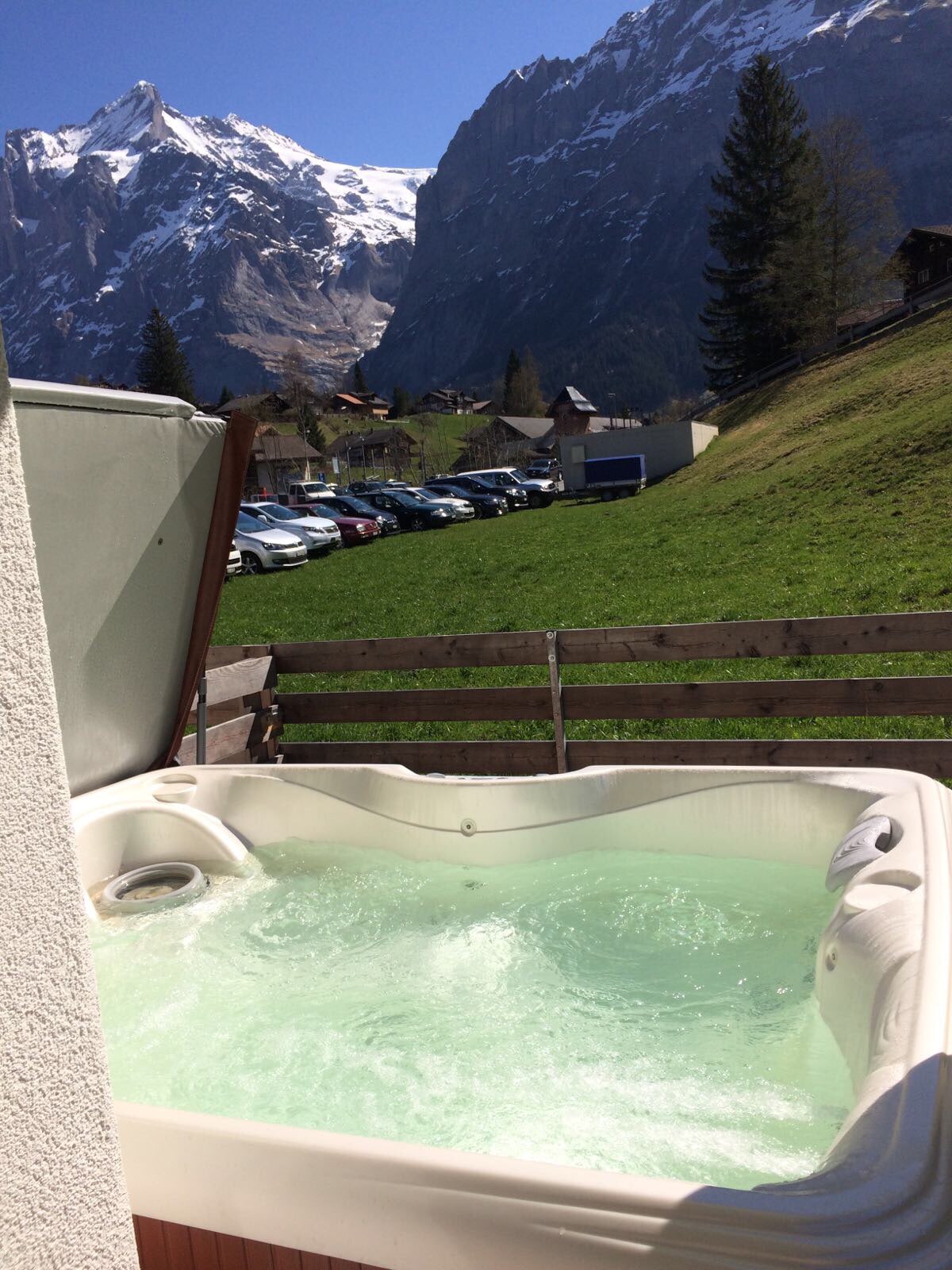 A private spa area features a bubbling hot tub, surrounded by wooden fencing. In the background, towering mountains are visible, capped with snow, under a clear blue sky. The lush green landscape adds to the serene setting.