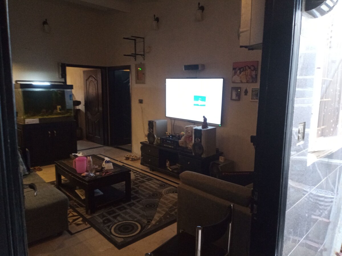 An open living area is visible, featuring a comfortable seating arrangement and a dark wooden television console. A wall-mounted TV displays content, while an aquarium is positioned in the corner. Natural light filters through partially open curtains, illuminating the space.