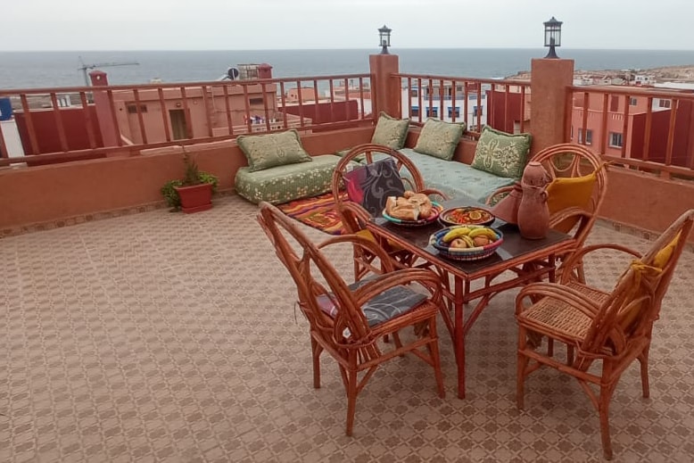 An outdoor terrace overlooks the sea, featuring a woven table surrounded by four rattan chairs. Colorful cushions are arranged on a lounge area, and a small potted plant adds a touch of greenery. A variety of fruits and pastries are displayed on the table, inviting relaxation.