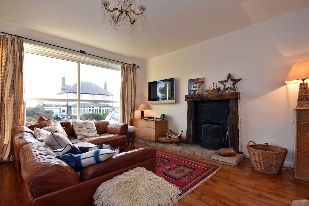 Spacious living area with wall mounted television and log burning fire. Front view towards fields and beach.