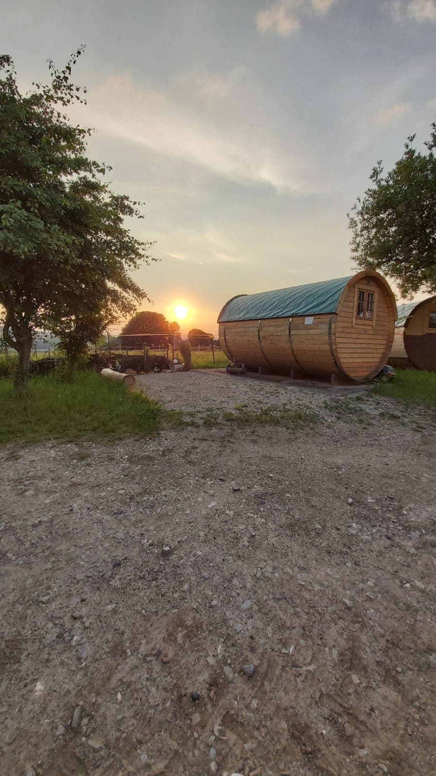 The image shows a wooden barrel-shaped accommodation set against a sunset backdrop, highlighting the warm colors of the sky. Surrounding greenery and a gravel path enhance the natural setting. The structure is partially illuminated, creating a welcoming atmosphere.