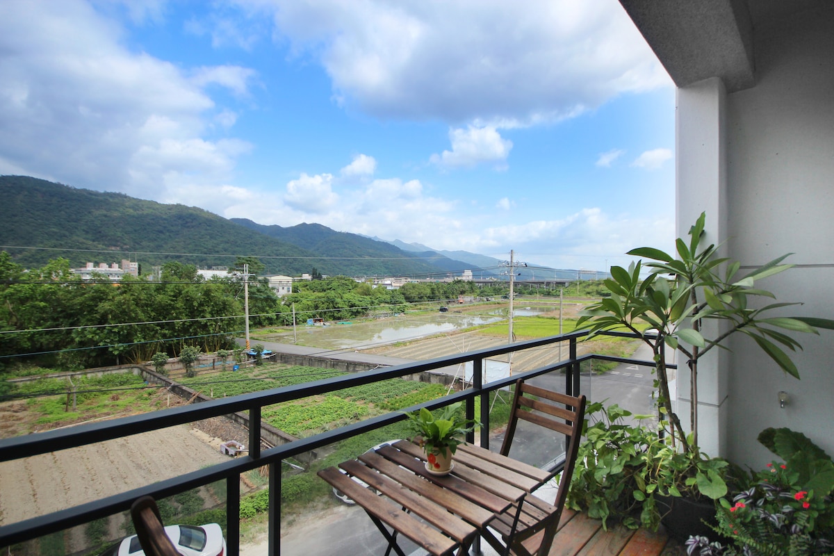 A balcony view is presented, featuring a wooden table and chairs surrounded by potted plants. In the background, rolling mountains and expansive fields can be seen under a partly cloudy sky, creating a serene landscape.