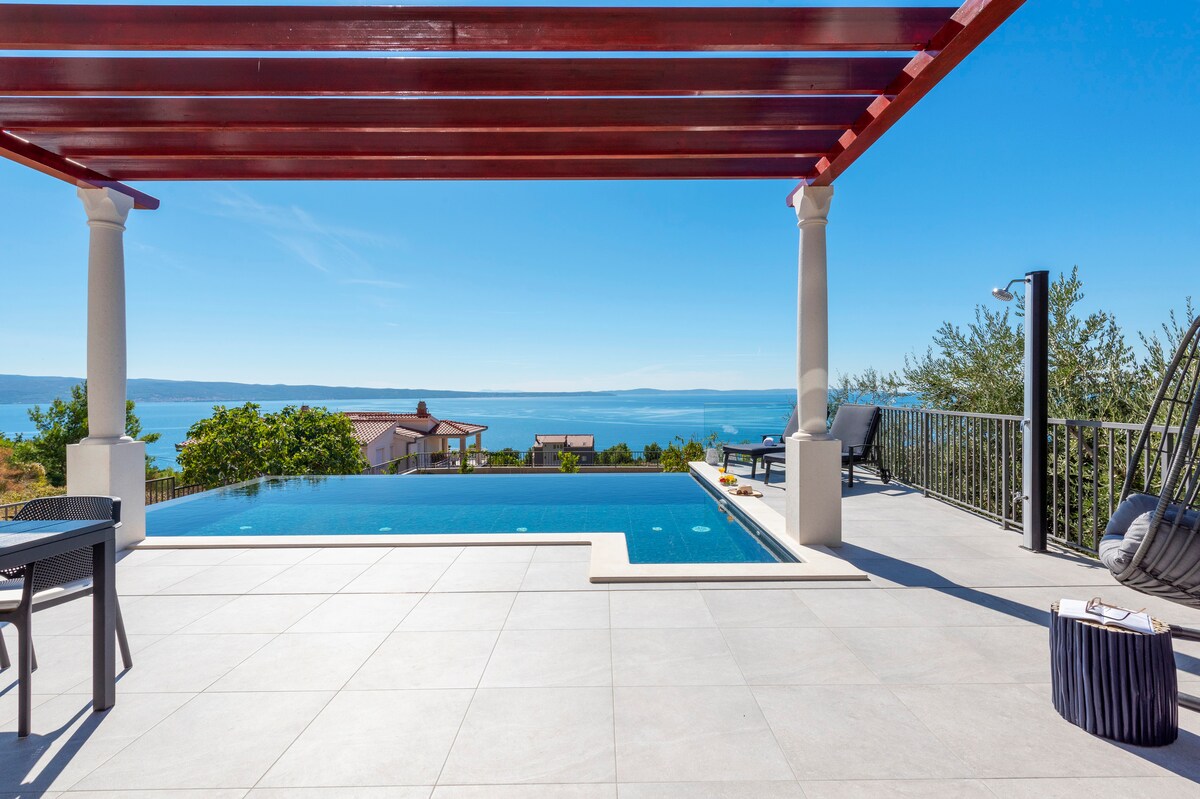 An outdoor area features a shimmering infinity pool with a hydromassage section, framed by a red pergola. A spacious patio offers views of the Adriatic Sea, complemented by a comfortable seating arrangement and a side table. Lush greenery is present in the background.