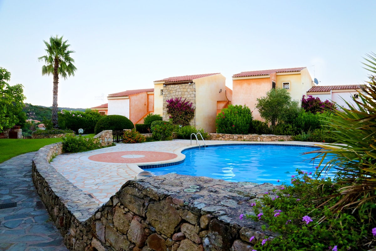 A clear view of the shared pool surrounded by well-maintained gardens is showcased. A stone wall encircles the pool area, while vibrant greenery and flowering plants provide a natural backdrop. The charming buildings of the complex are visible in the distance.